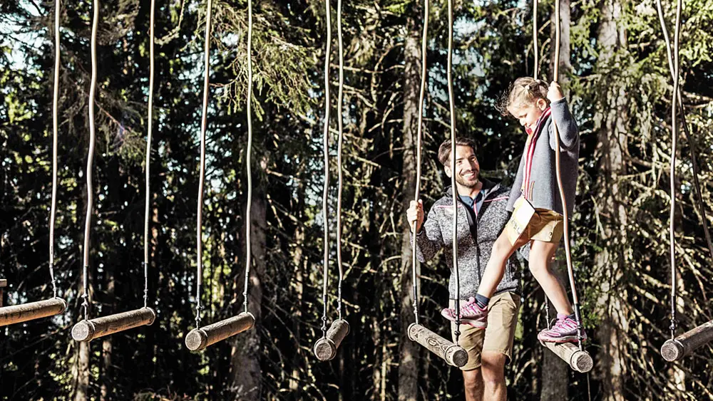 Vater hilft Tochter beim Balancieren an einer Station des Erlebniswanderwegs Schmidolins Feuertaufe auf der Schmittenhöhe