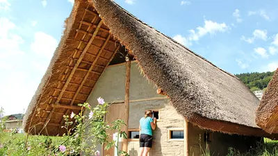 Frau sieht durch ein Fenster eines Hauses der Pfahlbausiedlung Wauwilermoos